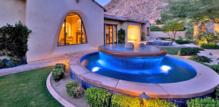 An elegant outdoor patio at dusk with a round blue-lit pool, stone house facade, palm trees, and a mountain backdrop, rendered in deep greens and twilight blues.