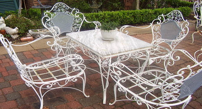 A white wrought-iron patio table and four chairs set on a brick patio, with a small potted plant on the table and green lawn beyond, in soft cream and sage tones.